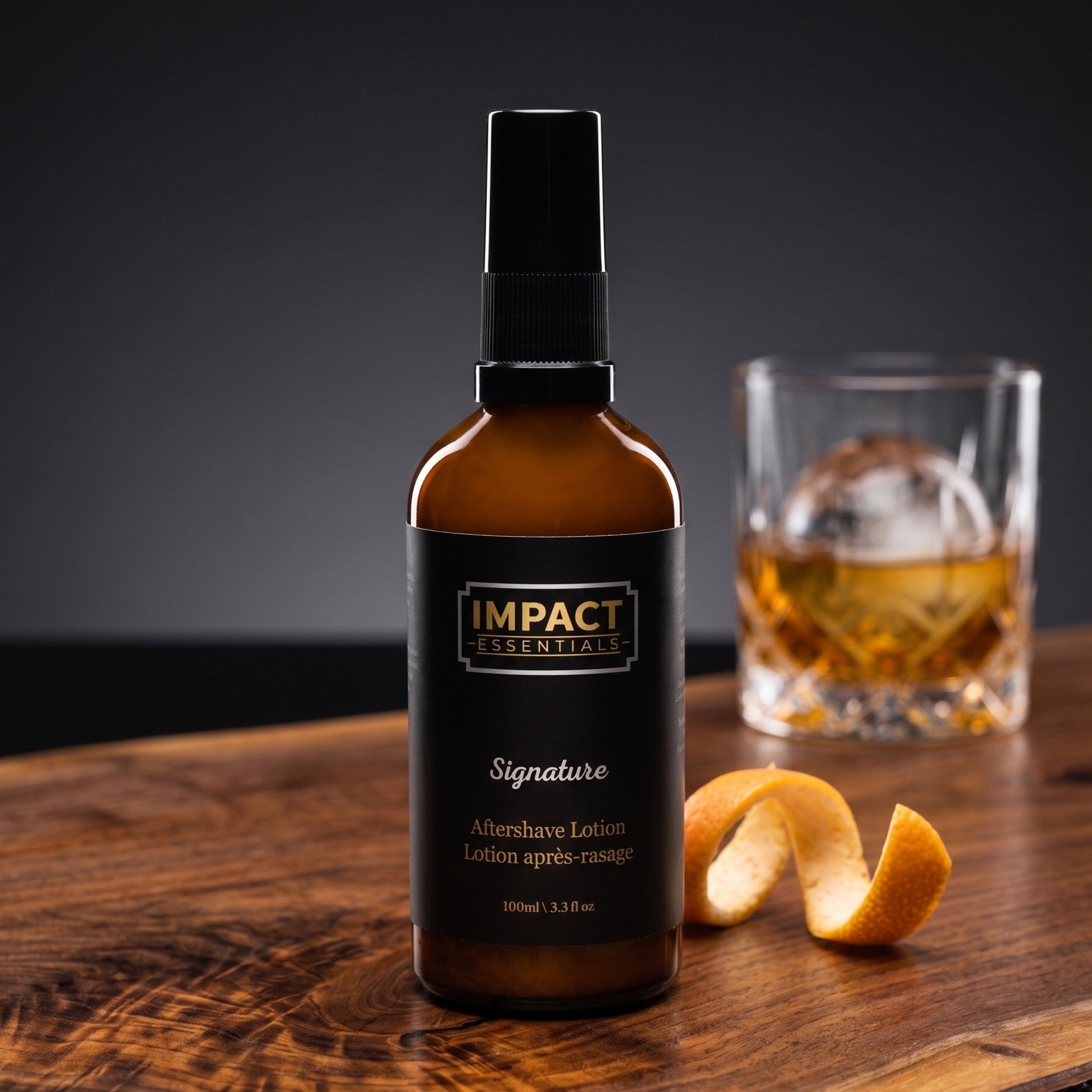 An amber glass bottle of Impact Essentials Signature Aftershave Lotion stands on a wooden table, with a blurred whiskey glass and a curled orange peel in the background.