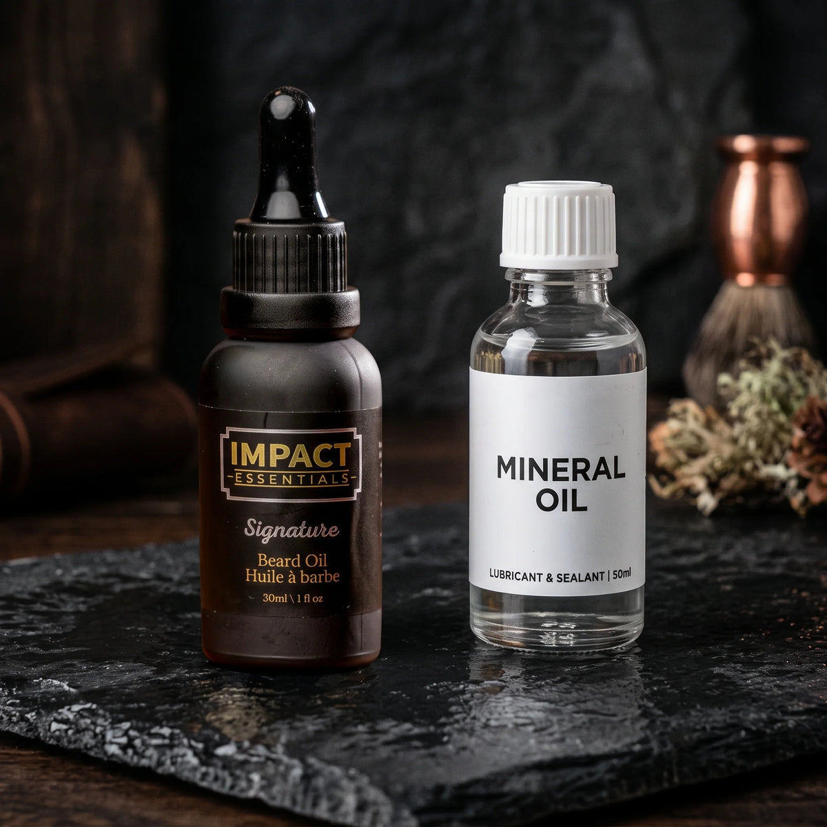 Bottle of IMPACT Essentials Signature natural botanical beard oil beside a generic bottle of mineral oil on a dark slate surface.