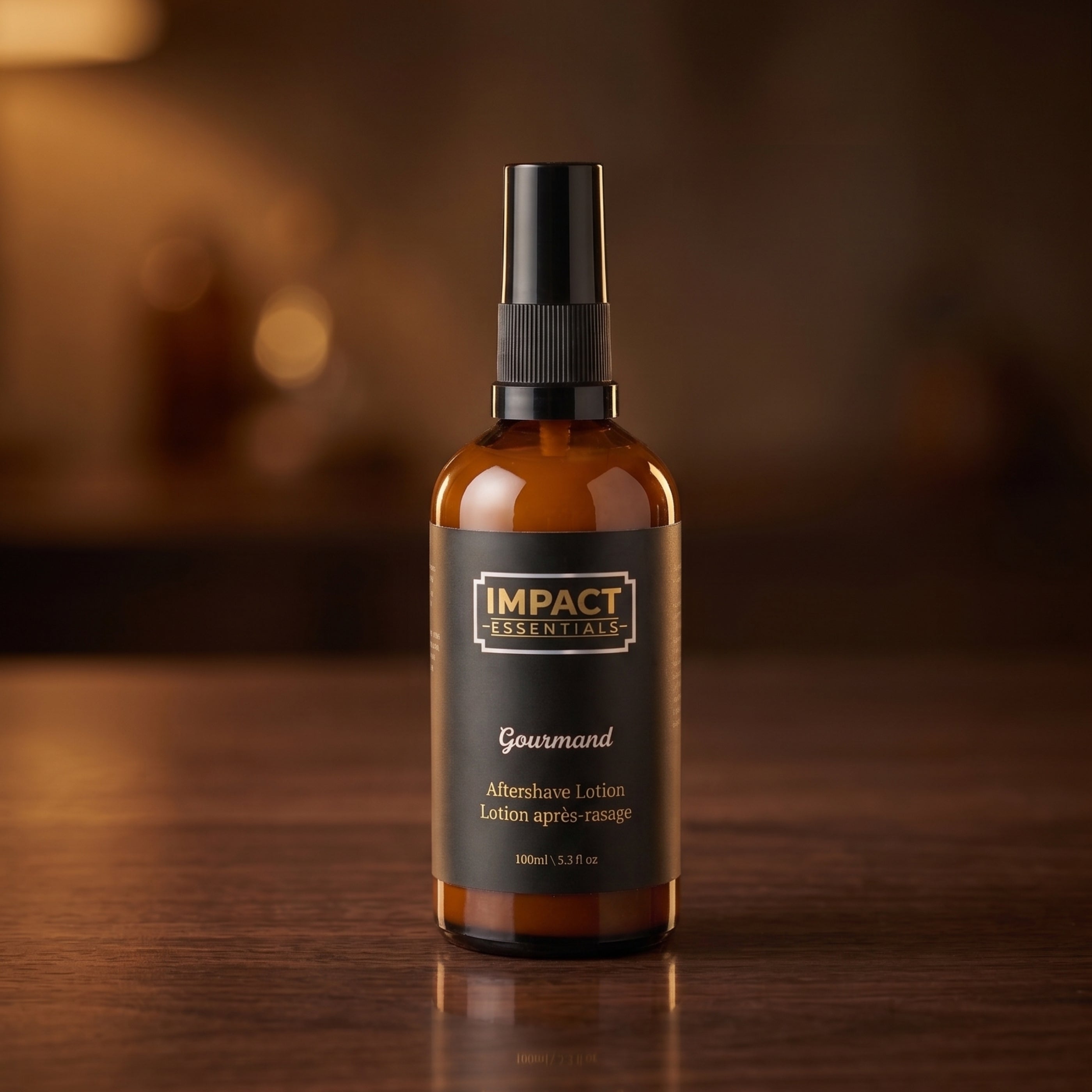 An amber glass spray bottle with a black spray top and a black label reading 'IMPACT ESSENTIALS' and 'Gourmand', placed on a dark wooden surface with a softly blurred warm brown background.