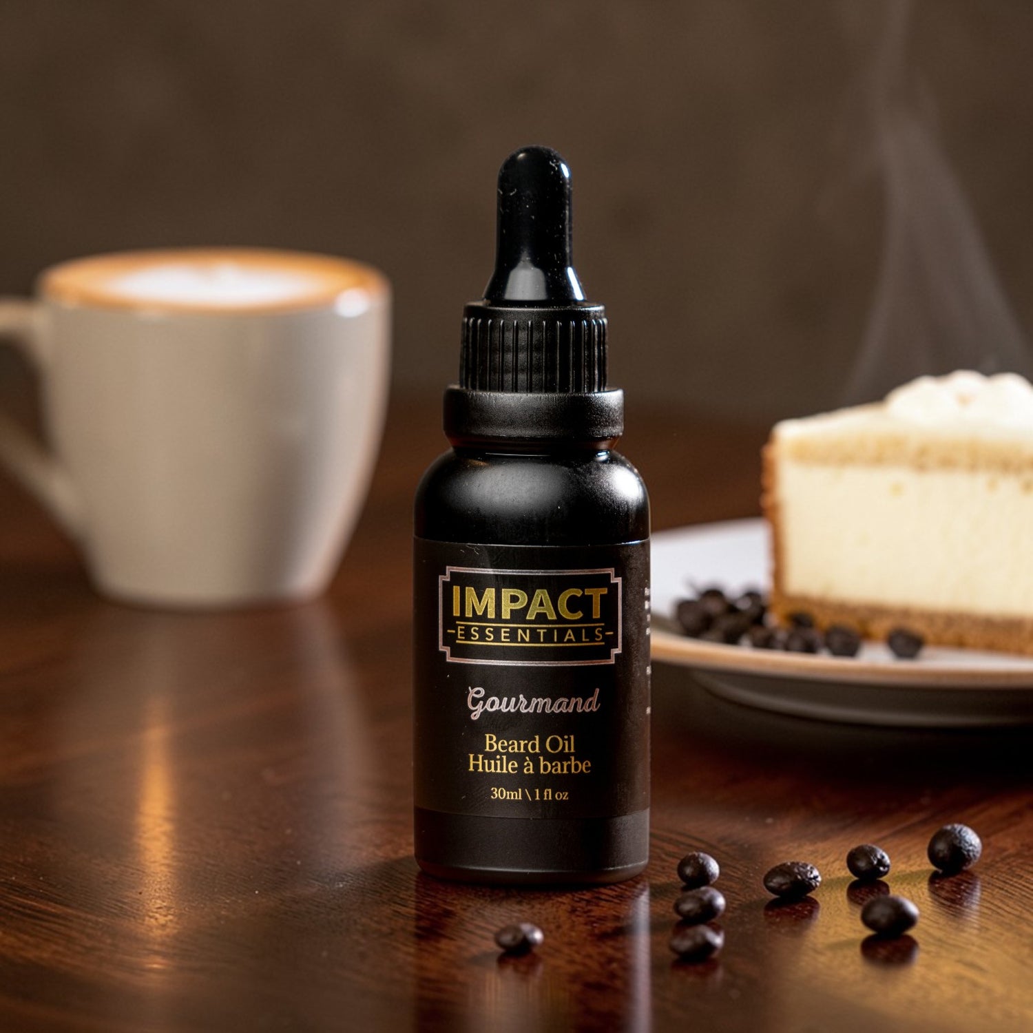 Bottle of Impact Essentials Gourmand Beard Oil on a wooden surface with a cup of coffee and cake in the background.