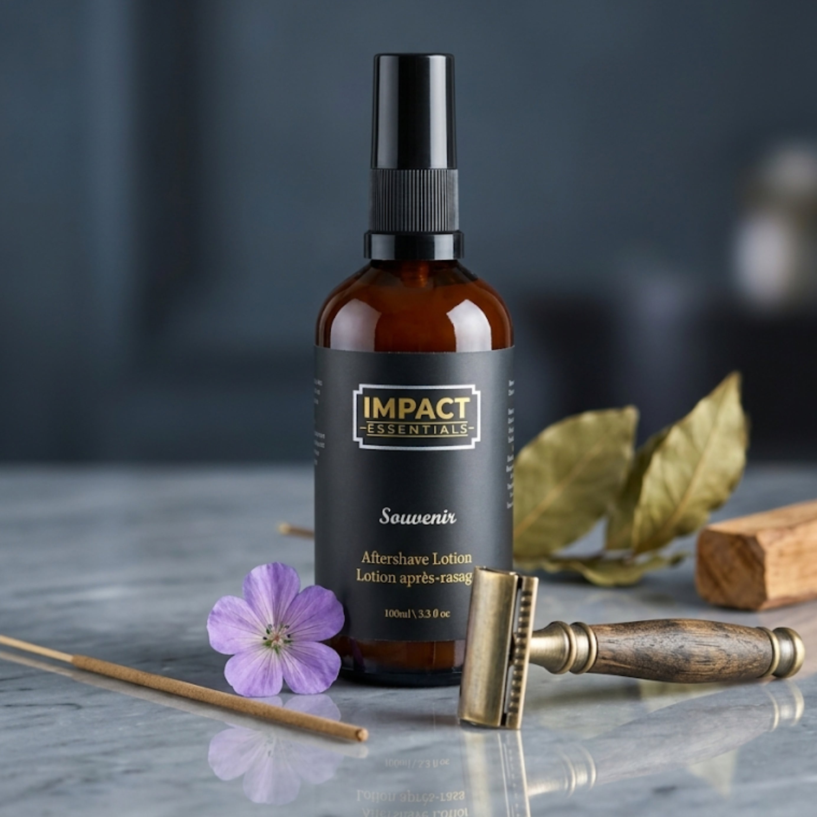 An amber glass spray bottle labeled IMPACT ESSENTIALS Souvenir Aftershave Lotion stands on a marble-like surface with a purple flower, brass razor, wooden stick, and dried leaves as styled props against a dark blurred background.